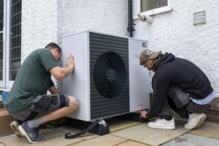 Two installers crouch next to a large heat pump, installing it to the side of a house.